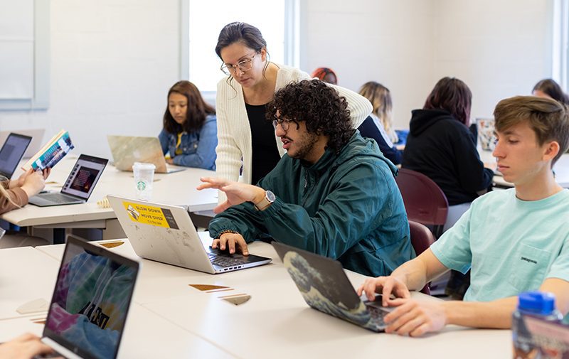 Professor helping a student while looking at a laptop surrounded by other student in the classroom