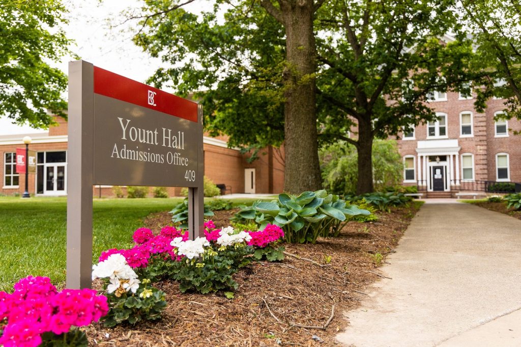 a sign among a row of flowers along a sidewalk that reads Yount Hall Admissions Office